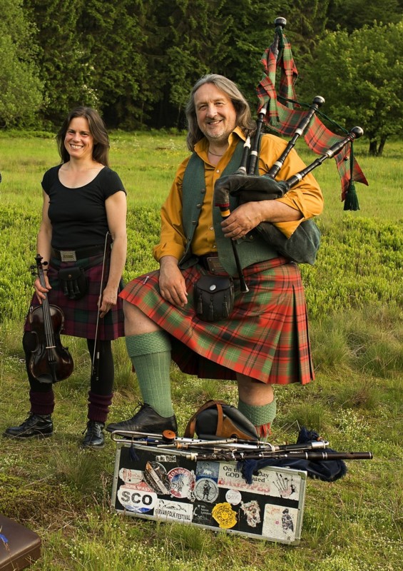 Johanna Wildhack trägt einen Mini-Kilt mit Sporran und hält ihre Geige, Quest trägt Kilt und Zubehör und hält einen großen Highland-Dudelsack. Auf einem Instrumentenkoffer vor ihm liegen ein irischer Dudelsack, eine Holzquerflöte, eine Low-Whistle und eine Tin-Whistle.